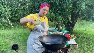 Önlüklü güzel bir kadın açık havada yemek hazırlıyor. Izgarada ateş yakıyor. Rahat bir açık hava ortamında malzemelerle ustalıkla uğraşırken sıcak havanın tadını çıkarıyor..