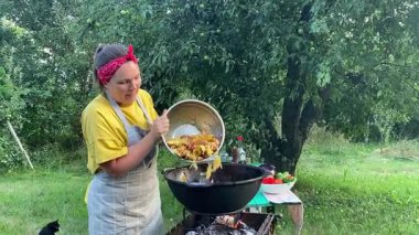 Önlüklü güzel bir kadın açık havada yemek hazırlıyor. Izgarada ateş yakıyor. Rahat bir açık hava ortamında malzemelerle ustalıkla uğraşırken sıcak havanın tadını çıkarıyor..