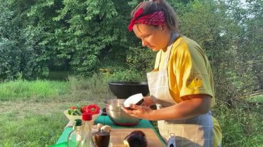 Önlüklü güzel bir kadın açık havada yemek hazırlıyor. Izgarada ateş yakıyor. Rahat bir açık hava ortamında malzemelerle ustalıkla uğraşırken sıcak havanın tadını çıkarıyor..