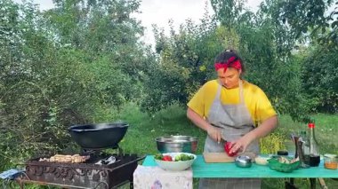Önlüklü güzel bir kadın açık havada yemek hazırlıyor. Izgarada ateş yakıyor. Rahat bir açık hava ortamında malzemelerle ustalıkla uğraşırken sıcak havanın tadını çıkarıyor..