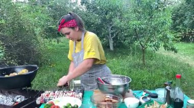 Önlüklü güzel bir kadın açık havada yemek hazırlıyor. Izgarada ateş yakıyor. Rahat bir açık hava ortamında malzemelerle ustalıkla uğraşırken sıcak havanın tadını çıkarıyor..