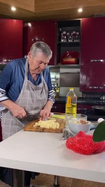 Modern bir ev mutfağında yemek hazırlayan önlüklü yaşlı bir kadının dikey hali. İçeriğiyle dikkatlice ilgilenir, ev ortamında deneyimli bir şekilde yemek pişirme sürecine odaklanır.