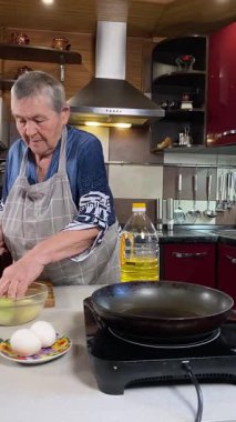 Modern bir ev mutfağında yemek hazırlayan önlüklü yaşlı bir kadının dikey hali. İçeriğiyle dikkatlice ilgilenir, ev ortamında deneyimli bir şekilde yemek pişirme sürecine odaklanır.