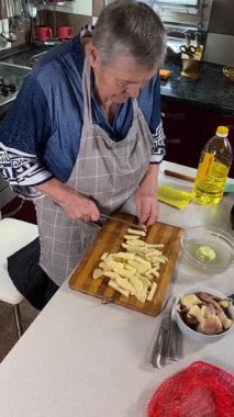 Modern bir ev mutfağında yemek hazırlayan önlüklü yaşlı bir kadının dikey hali. İçeriğiyle dikkatlice ilgilenir, ev ortamında deneyimli bir şekilde yemek pişirme sürecine odaklanır.