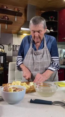 Modern bir ev mutfağında yemek hazırlayan önlüklü yaşlı bir kadının dikey hali. İçeriğiyle dikkatlice ilgilenir, ev ortamında deneyimli bir şekilde yemek pişirme sürecine odaklanır.