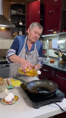 Modern bir ev mutfağında yemek hazırlayan önlüklü yaşlı bir kadının dikey hali. İçeriğiyle dikkatlice ilgilenir, ev ortamında deneyimli bir şekilde yemek pişirme sürecine odaklanır.