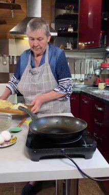 Modern bir ev mutfağında yemek hazırlayan önlüklü yaşlı bir kadının dikey hali. İçeriğiyle dikkatlice ilgilenir, ev ortamında deneyimli bir şekilde yemek pişirme sürecine odaklanır.