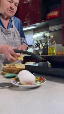 Modern bir ev mutfağında yemek hazırlayan önlüklü yaşlı bir kadının dikey hali. İçeriğiyle dikkatlice ilgilenir, ev ortamında deneyimli bir şekilde yemek pişirme sürecine odaklanır.