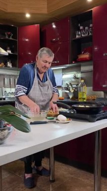 Modern bir ev mutfağında yemek hazırlayan önlüklü yaşlı bir kadının dikey hali. İçeriğiyle dikkatlice ilgilenir, ev ortamında deneyimli bir şekilde yemek pişirme sürecine odaklanır.