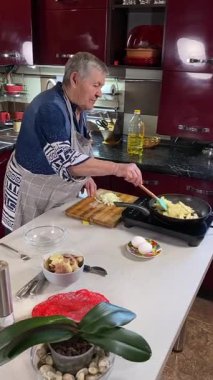 Modern bir ev mutfağında yemek hazırlayan önlüklü yaşlı bir kadının dikey hali. İçeriğiyle dikkatlice ilgilenir, ev ortamında deneyimli bir şekilde yemek pişirme sürecine odaklanır.