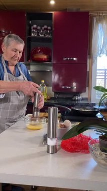 Modern bir ev mutfağında yemek hazırlayan önlüklü yaşlı bir kadının dikey hali. İçeriğiyle dikkatlice ilgilenir, ev ortamında deneyimli bir şekilde yemek pişirme sürecine odaklanır.