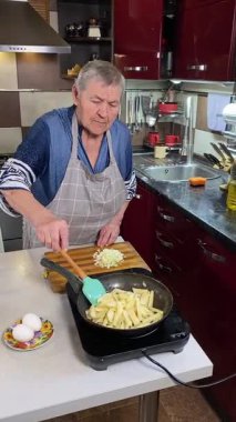 Modern bir ev mutfağında yemek hazırlayan önlüklü yaşlı bir kadının dikey hali. İçeriğiyle dikkatlice ilgilenir, ev ortamında deneyimli bir şekilde yemek pişirme sürecine odaklanır.