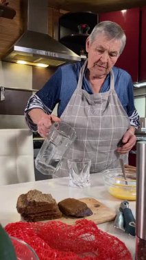 Modern bir ev mutfağında yemek hazırlayan önlüklü yaşlı bir kadının dikey hali. İçeriğiyle dikkatlice ilgilenir, ev ortamında deneyimli bir şekilde yemek pişirme sürecine odaklanır.