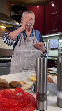 Modern bir ev mutfağında yemek hazırlayan önlüklü yaşlı bir kadının dikey hali. İçeriğiyle dikkatlice ilgilenir, ev ortamında deneyimli bir şekilde yemek pişirme sürecine odaklanır.