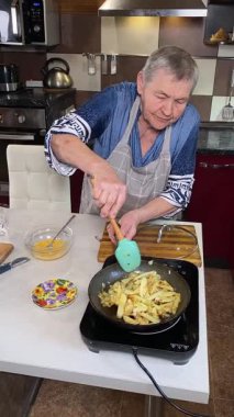 Modern bir ev mutfağında yemek hazırlayan önlüklü yaşlı bir kadının dikey hali. İçeriğiyle dikkatlice ilgilenir, ev ortamında deneyimli bir şekilde yemek pişirme sürecine odaklanır.