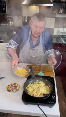 Modern bir ev mutfağında yemek hazırlayan önlüklü yaşlı bir kadının dikey hali. İçeriğiyle dikkatlice ilgilenir, ev ortamında deneyimli bir şekilde yemek pişirme sürecine odaklanır.