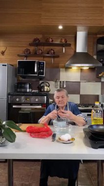 Modern bir ev mutfağında yemek hazırlayan önlüklü yaşlı bir kadının dikey hali. İçeriğiyle dikkatlice ilgilenir, ev ortamında deneyimli bir şekilde yemek pişirme sürecine odaklanır.