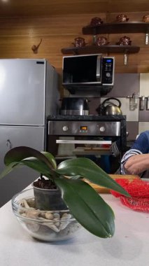 Modern bir ev mutfağında yemek hazırlayan önlüklü yaşlı bir kadının dikey hali. İçeriğiyle dikkatlice ilgilenir, ev ortamında deneyimli bir şekilde yemek pişirme sürecine odaklanır.