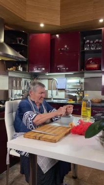 Modern bir ev mutfağında yemek hazırlayan önlüklü yaşlı bir kadının dikey hali. İçeriğiyle dikkatlice ilgilenir, ev ortamında deneyimli bir şekilde yemek pişirme sürecine odaklanır.