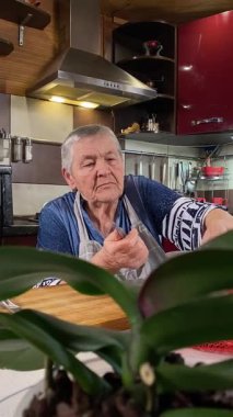 Modern bir ev mutfağında yemek hazırlayan önlüklü yaşlı bir kadının dikey hali. İçeriğiyle dikkatlice ilgilenir, ev ortamında deneyimli bir şekilde yemek pişirme sürecine odaklanır.