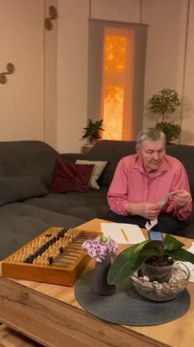 Oturma odasında vergileri, elektrik faturalarını akıllı telefon ve abaküs kullanarak hesap yapan yaşlı bir kadın. Odaklanmış ve düşünceli. Ekonomisini hem modern hem de geleneksel araçlarla yönetiyor.