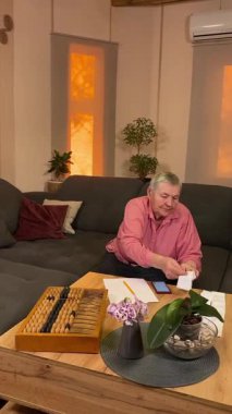 Oturma odasında vergileri, elektrik faturalarını akıllı telefon ve abaküs kullanarak hesap yapan yaşlı bir kadın. Odaklanmış ve düşünceli. Ekonomisini hem modern hem de geleneksel araçlarla yönetiyor.