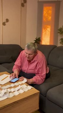 Oturma odasında vergileri, elektrik faturalarını akıllı telefon ve abaküs kullanarak hesap yapan yaşlı bir kadın. Odaklanmış ve düşünceli. Ekonomisini hem modern hem de geleneksel araçlarla yönetiyor.