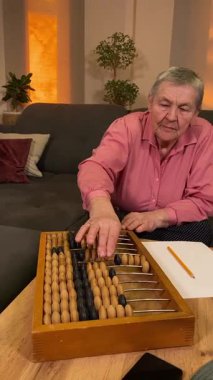 Oturma odasında vergileri, elektrik faturalarını akıllı telefon ve abaküs kullanarak hesap yapan yaşlı bir kadın. Odaklanmış ve düşünceli. Ekonomisini hem modern hem de geleneksel araçlarla yönetiyor.