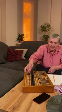 Oturma odasında vergileri, elektrik faturalarını akıllı telefon ve abaküs kullanarak hesap yapan yaşlı bir kadın. Odaklanmış ve düşünceli. Ekonomisini hem modern hem de geleneksel araçlarla yönetiyor.