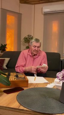 Oturma odasında vergileri, elektrik faturalarını akıllı telefon ve abaküs kullanarak hesap yapan yaşlı bir kadın. Odaklanmış ve düşünceli. Ekonomisini hem modern hem de geleneksel araçlarla yönetiyor.