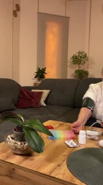 Sıcaklığı, oturma odasındaki kan basıncını ölçerek sağlıkla ilgilenen dikey yaşlı kadın. Günlük ilaç organizatörü ve ilaç çevrede görünüyor, rutin ve tıbbi bakımını yansıtıyor.