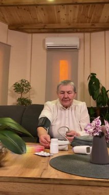 Sıcaklığı, oturma odasındaki kan basıncını ölçerek sağlıkla ilgilenen dikey yaşlı kadın. Günlük ilaç organizatörü ve ilaç çevrede görünüyor, rutin ve tıbbi bakımını yansıtıyor.