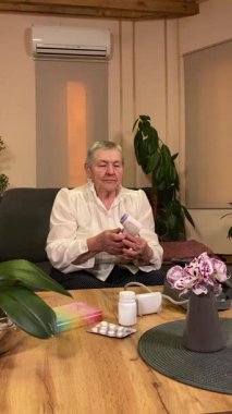 Sıcaklığı, oturma odasındaki kan basıncını ölçerek sağlıkla ilgilenen dikey yaşlı kadın. Günlük ilaç organizatörü ve ilaç çevrede görünüyor, rutin ve tıbbi bakımını yansıtıyor.