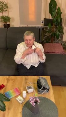 Sıcaklığı, oturma odasındaki kan basıncını ölçerek sağlıkla ilgilenen dikey yaşlı kadın. Günlük ilaç organizatörü ve ilaç çevrede görünüyor, rutin ve tıbbi bakımını yansıtıyor.