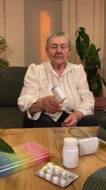 Sıcaklığı, oturma odasındaki kan basıncını ölçerek sağlıkla ilgilenen dikey yaşlı kadın. Günlük ilaç organizatörü ve ilaç çevrede görünüyor, rutin ve tıbbi bakımını yansıtıyor.