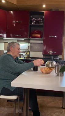 Dikey yaşlı kadın sıcak bir ev mutfağında öğle yemeği yiyor, sıcak bir ev yemeği yiyor. Yavaş ve dikkatli bir şekilde yiyor, tanıdık yiyeceklerin rahatlığını ve etrafındaki sakin atmosferi takdir ediyor.. 