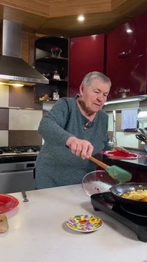 Dikey yaşlı kadın sıcak bir ev mutfağında öğle yemeği yiyor, sıcak bir ev yemeği yiyor. Yavaş ve dikkatli bir şekilde yiyor, tanıdık yiyeceklerin rahatlığını ve etrafındaki sakin atmosferi takdir ediyor.. 