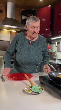 Dikey yaşlı kadın sıcak bir ev mutfağında öğle yemeği yiyor, sıcak bir ev yemeği yiyor. Yavaş ve dikkatli bir şekilde yiyor, tanıdık yiyeceklerin rahatlığını ve etrafındaki sakin atmosferi takdir ediyor.. 