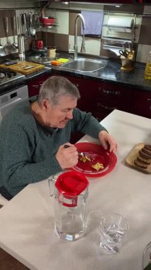 Dikey yaşlı kadın sıcak bir ev mutfağında öğle yemeği yiyor, sıcak bir ev yemeği yiyor. Yavaş ve dikkatli bir şekilde yiyor, tanıdık yiyeceklerin rahatlığını ve etrafındaki sakin atmosferi takdir ediyor.. 