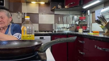 Modern bir ev mutfağında yemek hazırlayan kısa gri saçlı yaşlı bir kadın. İçeriğiyle dikkatlice ilgilenir, ev ortamında deneyimli bir şekilde yemek pişirme sürecine odaklanır. 