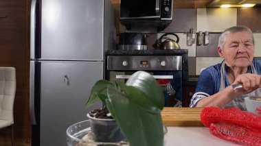 Modern bir ev mutfağında yemek hazırlayan kısa gri saçlı yaşlı bir kadın. İçeriğiyle dikkatlice ilgilenir, ev ortamında deneyimli bir şekilde yemek pişirme sürecine odaklanır. 