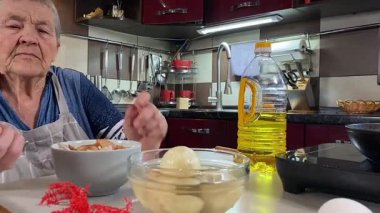 Modern bir ev mutfağında yemek hazırlayan kısa gri saçlı yaşlı bir kadın. İçeriğiyle dikkatlice ilgilenir, ev ortamında deneyimli bir şekilde yemek pişirme sürecine odaklanır. 