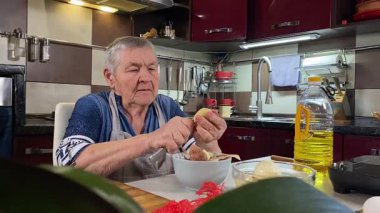 Modern bir ev mutfağında yemek hazırlayan kısa gri saçlı yaşlı bir kadın. İçeriğiyle dikkatlice ilgilenir, ev ortamında deneyimli bir şekilde yemek pişirme sürecine odaklanır. 