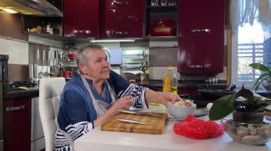 Modern bir ev mutfağında yemek hazırlayan kısa gri saçlı yaşlı bir kadın. İçeriğiyle dikkatlice ilgilenir, ev ortamında deneyimli bir şekilde yemek pişirme sürecine odaklanır. 