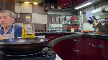 Modern bir ev mutfağında yemek hazırlayan kısa gri saçlı yaşlı bir kadın. İçeriğiyle dikkatlice ilgilenir, ev ortamında deneyimli bir şekilde yemek pişirme sürecine odaklanır. 