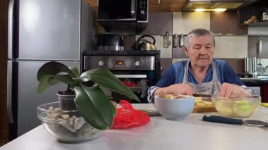 Modern bir ev mutfağında yemek hazırlayan kısa gri saçlı yaşlı bir kadın. İçeriğiyle dikkatlice ilgilenir, ev ortamında deneyimli bir şekilde yemek pişirme sürecine odaklanır. 