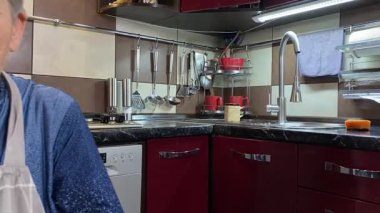 Modern bir ev mutfağında yemek hazırlayan kısa gri saçlı yaşlı bir kadın. İçeriğiyle dikkatlice ilgilenir, ev ortamında deneyimli bir şekilde yemek pişirme sürecine odaklanır. 