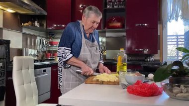 Modern bir ev mutfağında yemek hazırlayan kısa gri saçlı yaşlı bir kadın. İçeriğiyle dikkatlice ilgilenir, ev ortamında deneyimli bir şekilde yemek pişirme sürecine odaklanır. 