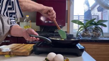 Modern bir ev mutfağında yemek hazırlayan kısa gri saçlı yaşlı bir kadın. İçeriğiyle dikkatlice ilgilenir, ev ortamında deneyimli bir şekilde yemek pişirme sürecine odaklanır. 