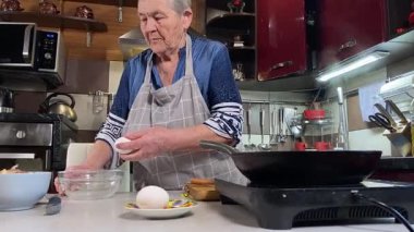 Modern bir ev mutfağında yemek hazırlayan kısa gri saçlı yaşlı bir kadın. İçeriğiyle dikkatlice ilgilenir, ev ortamında deneyimli bir şekilde yemek pişirme sürecine odaklanır. 